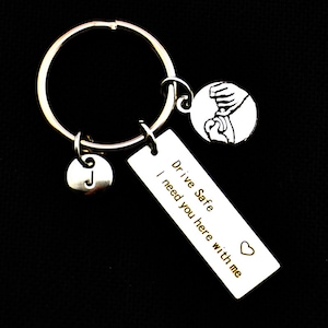 Puede incluir: Llavero plateado con un anillo grande, una etiqueta rectangular con el texto "Drive Safe I need you here with me", un dije redondo con una mano sosteniendo un volante y un dije redondo con la letra "J".