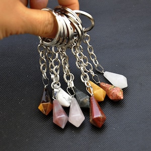 May include: A collection of gemstone keychains with silver-colored chains and rings. Each keychain features a different colored, faceted gemstone pendant, including brown, white, pink, and red. The keychains are held against a dark background.