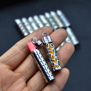 Op de afbeelding: Twee zilveren metalen hangerbehangers met verschillende ontwerpen. Een hanger is open en toont een roze papiertje erin. De andere hanger heeft een geel bloemenontwerp.