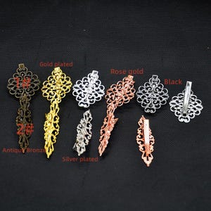 May include: An assortment of decorative hair clips in antique bronze, gold, silver, rose gold, and black finishes. Each clip has an elaborate filigree pattern. Text labels identify each colour. The clips are displayed against a dark background.