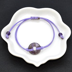 May include: A purple string bracelet with a round, purple stone charm and two small purple beads. The bracelet is tied with a knot and is on a white plate.