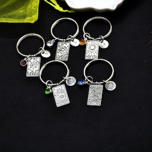 Puede incluir: Llaveros plateados con colgantes de cartas del tarot, cada uno con un diseño de carta diferente y una pequeña piedra preciosa. Los llaveros también incluyen un pequeño dije inicial. Las cartas del tarot incluyen "El Mundo", "La Luna", "El Sol", "Fortuna" y "La Estrella".