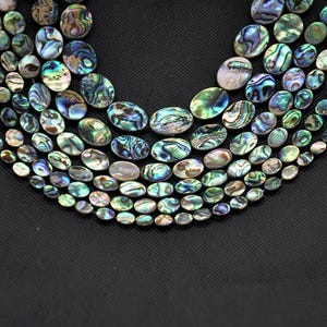 May include: A collection of iridescent abalone shell beads in various sizes and shapes. The oval-shaped beads display a range of colors, including blues, greens, and purples, with unique patterns. The beads are arranged on a black surface.