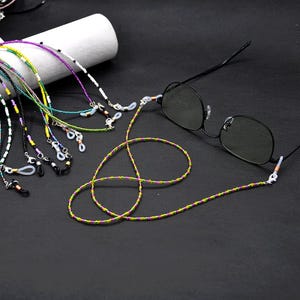 May include: A selection of beaded eyeglass chains in various colours, including green, purple, and yellow. One pair of black-framed glasses is attached to a chain. The chains are designed to hold eyeglasses.