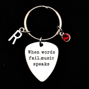 Könnte beinhalten: Silberner Gitarren-Plektrum-Schlüsselanhänger mit dem Text "When words fail. music speaks" und einem roten Strass-Anhänger.