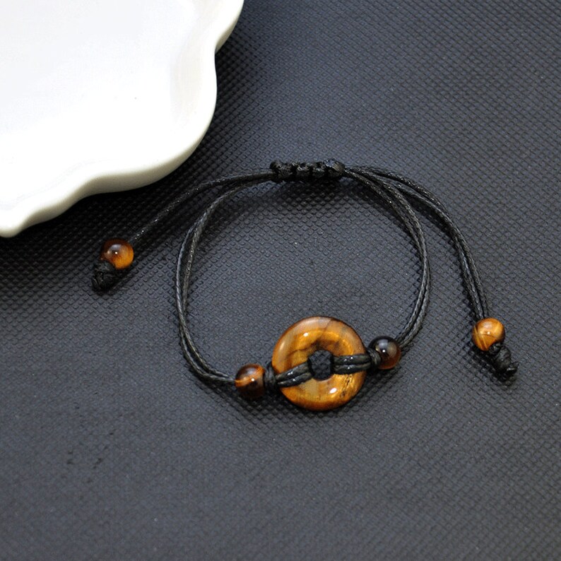 May include: A black string bracelet with a brown tiger's eye stone charm in the shape of a circle. The bracelet has two smaller brown tiger's eye beads on either side of the charm.