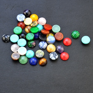 May include: Assortment of colorful cabochons, including turquoise, green, red, blue, and brown, in various shapes and sizes. The cabochons are arranged on a black surface.