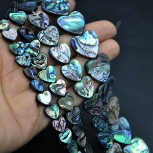 Könnte beinhalten: Eine Sammlung herzförmiger Perlen mit schillernden, abaloneartigen Oberflächen. Die Perlen zeigen eine Reihe von Farben, darunter Blau-, Grün- und Lilatöne, mit einem schimmernden, strukturierten Aussehen. Die Perlen sind aufgefädelt.