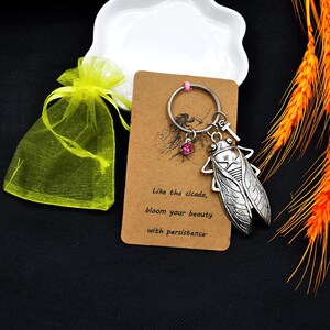 May include: A silver-colored cicada keychain with a pink gem charm, attached to a brown card with the text "Like the cicada, bloom your beauty with persistence." A yellow organza gift bag is also included.