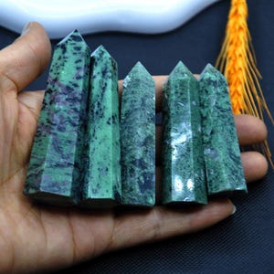 May include: Five green and purple gemstone obelisks with pointed tops. The obelisks are held in a hand, showcasing their polished surfaces and natural color variations. The gemstones have a marbled appearance.