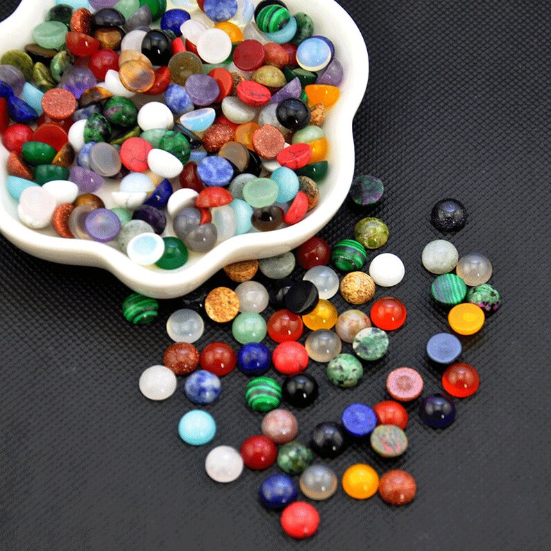 May include: A white bowl filled with a variety of colorful round gemstone cabochons. The bowl is tipped over, spilling the cabochons onto a black surface.