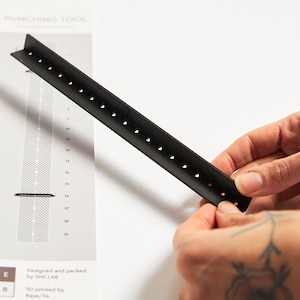 Könnte beinhalten: Ein schwarzes Plastiklineal mit Löchern am Rand. Das Lineal ist mit Zahlen von 10 bis 19 gekennzeichnet. Das Lineal wird von einer Hand mit einem Tattoo gehalten.