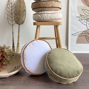 Pu&ograve; includere: Una pila di quattro cuscini rotondi di diversi colori e trame. I cuscini sono impilati su uno sgabello di legno. I cuscini sono in tonalit&agrave; di marrone, beige e bianco. Lo sgabello &egrave; in legno chiaro. C'&egrave; un cuscino verde sul pavimento davanti allo sgabello.