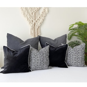 May include: Five decorative throw pillows on a white bed. Two pillows are gray with white piping. Two pillows are black and white with a geometric pattern and white piping. One pillow is black velvet with white piping.