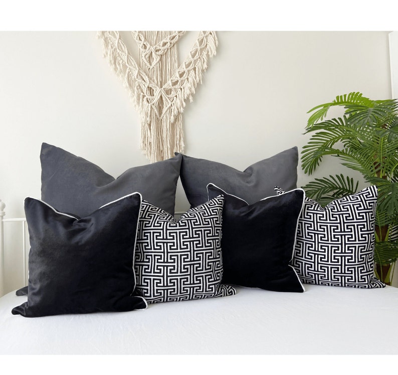 May include: Five decorative throw pillows on a white bed. Two pillows are gray with white piping. Two pillows are black and white with a geometric pattern and white piping. One pillow is black velvet with white piping.