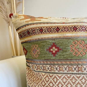 May include: A decorative throw pillow with a geometric pattern in shades of green, brown, and red. The pillow has a woven texture and a traditional design.