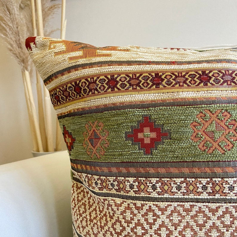 May include: A decorative throw pillow with a geometric pattern in shades of green, brown, and red. The pillow has a woven texture and a traditional design.