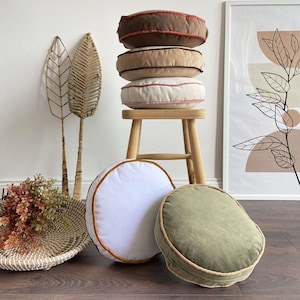 May include: A stack of four round cushions with different coloured fabric covers. The cushions are stacked on a wooden stool. The cushions are all different colours, including brown, beige, and white. The cushions have a decorative trim around the edges.