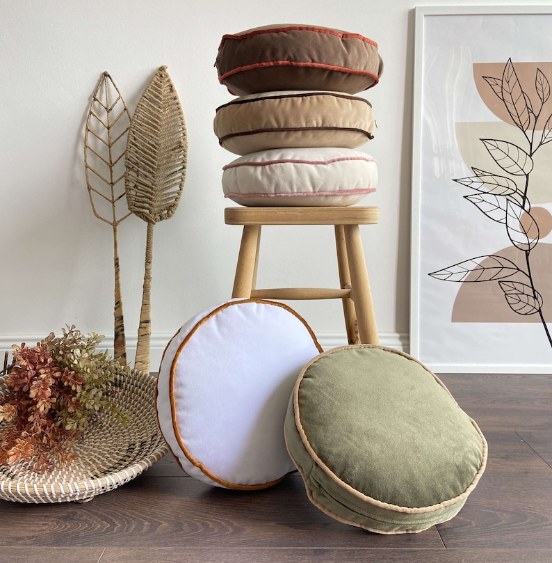 May include: A stack of four round cushions with different coloured fabric covers. The cushions are stacked on a wooden stool. The cushions are all different colours, including brown, beige, and white. The cushions have a decorative trim around the edges.