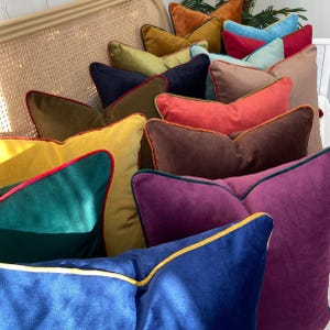 May include: A collection of decorative throw pillows in various colors and textures, including velvet, with contrasting trim. The pillows are arranged in a stack on a wicker chair.