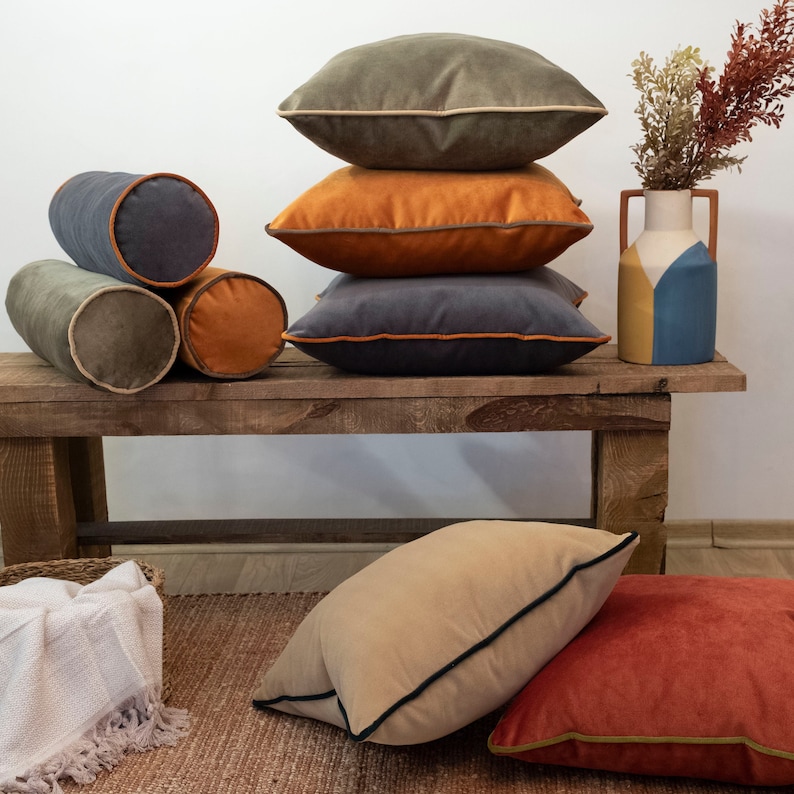 May include: A collection of decorative throw pillows in various colors and textures, including a light brown, dark brown, gray, and orange. The pillows are arranged on a wooden bench with a vase of dried flowers in the background.