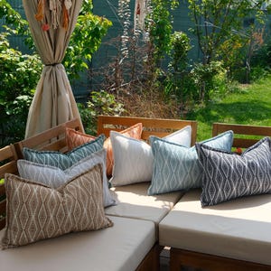 K&ouml;nnte beinhalten: Eine Auswahl an dekorativen Kissen in Beige, Wei&szlig;, T&uuml;rkis und Marineblau mit geometrischen Mustern. Die Kissen liegen auf Gartenm&ouml;beln aus Holz, umgeben von viel Gr&uuml;n.