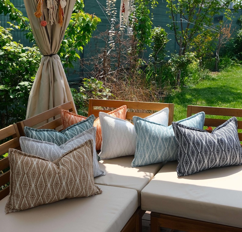 K&ouml;nnte beinhalten: Eine Auswahl an dekorativen Kissen in Beige, Wei&szlig;, T&uuml;rkis und Marineblau mit geometrischen Mustern. Die Kissen liegen auf Gartenm&ouml;beln aus Holz, umgeben von viel Gr&uuml;n.