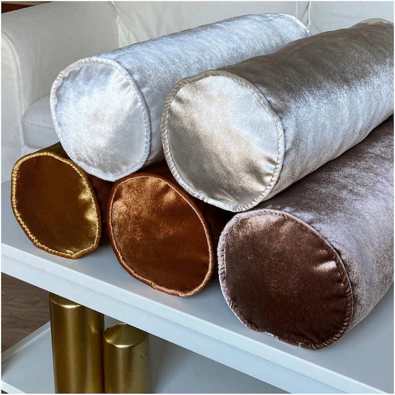 K&ouml;nnte beinhalten: F&uuml;nf zylindrische Kissen aus Samt in verschiedenen Farben, darunter Gold, Braun und zwei Graut&ouml;ne. Die Kissen sind auf einer wei&szlig;en Oberfl&auml;che angeordnet.