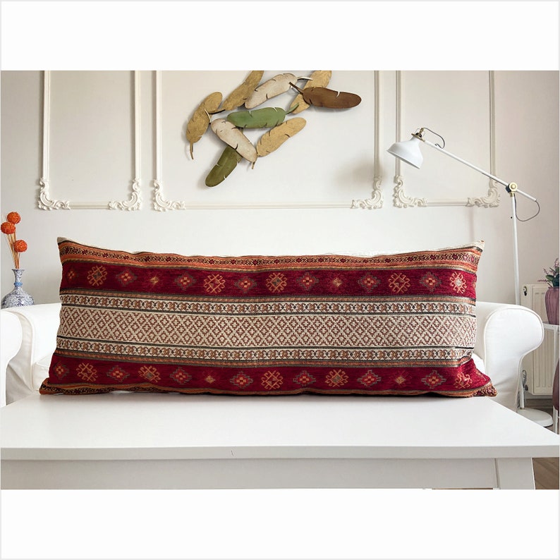 May include: A long, rectangular pillow with a red, white, and brown geometric pattern. The pillow is made of a soft, woven fabric and has a fringe trim.