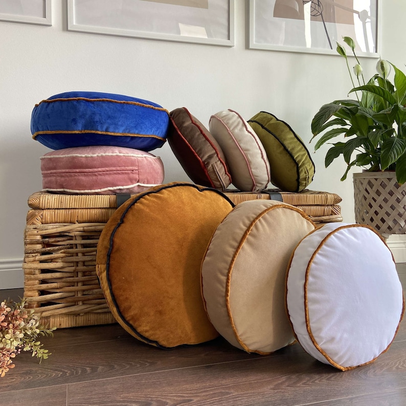 May include: A collection of round velvet cushions in various colours, including blue, pink, green, brown, beige, and white. The cushions have a contrasting trim around the edges and are arranged on a wicker basket.