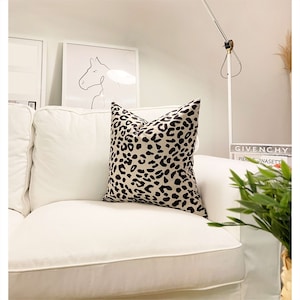 Puede incluir: Un sof&aacute; blanco con un coj&iacute;n de estampado de leopardo negro y beige. El sof&aacute; est&aacute; en una sala de estar con una l&aacute;mpara blanca y un libro con el texto "GIVENCHY PIERRE DONASETTI" en una mesa.