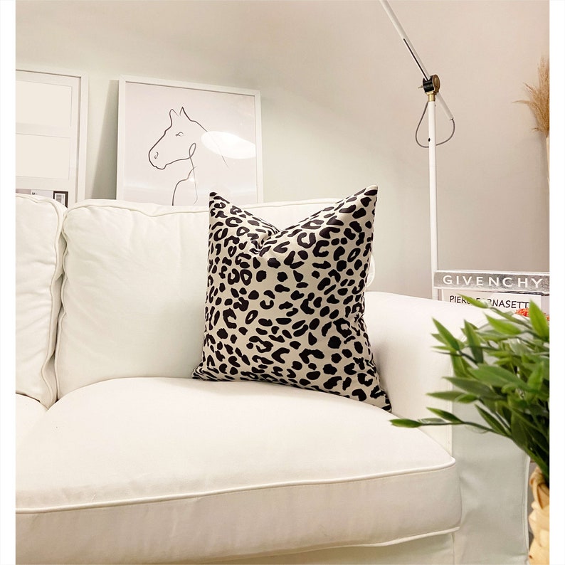 Puede incluir: Un sof&aacute; blanco con un coj&iacute;n de estampado de leopardo negro y beige. El sof&aacute; est&aacute; en una sala de estar con una l&aacute;mpara blanca y un libro con el texto "GIVENCHY PIERRE DONASETTI" en una mesa.