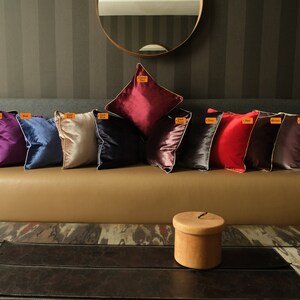 Puede incluir: Una fila de 10 cojines decorativos en varios colores, incluyendo negro, morado, azul, beige, azul oscuro, rojo, gris, burdeos y blanco. Los cojines est&aacute;n dispuestos en un sof&aacute; marr&oacute;n con una caja de madera delante del sof&aacute;.
