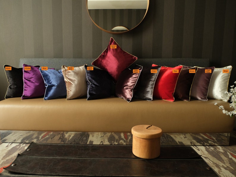 Puede incluir: Una fila de 10 cojines decorativos en varios colores, incluyendo negro, morado, azul, beige, azul oscuro, rojo, gris, burdeos y blanco. Los cojines est&aacute;n dispuestos en un sof&aacute; marr&oacute;n con una caja de madera delante del sof&aacute;.