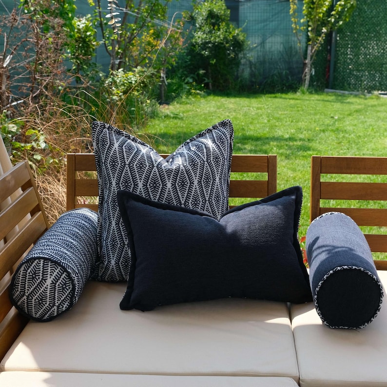 K&ouml;nnte beinhalten: Eine Auswahl an Outdoor-Kissen in verschiedenen Formen und Mustern. Enth&auml;lt ein gro&szlig;es quadratisches Kissen mit einem schwarz-wei&szlig;en geometrischen Design, ein rechteckiges schwarzes Kissen und zwei zylindrische Kissen mit &auml;hnlichen Mustern. Die Kissen sind auf einem cremefarbenen Outdoor-Sofa angeordnet.