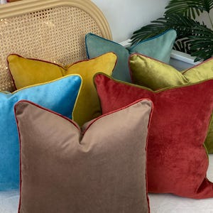 May include: A collection of six velvet throw pillows with contrasting colored piping. The pillows are in shades of mustard yellow, teal, olive green, brown, and red.