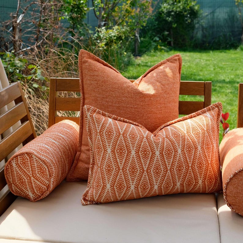 K&ouml;nnte beinhalten: Eine Auswahl an orangefarbenen Outdoor-Kissen in verschiedenen Formen. Die Kissen umfassen eine Rolle, quadratische und rechteckige Kissen. Einige haben ein wei&szlig;es geometrisches Muster. Die Kissen liegen auf einer Holzbank.