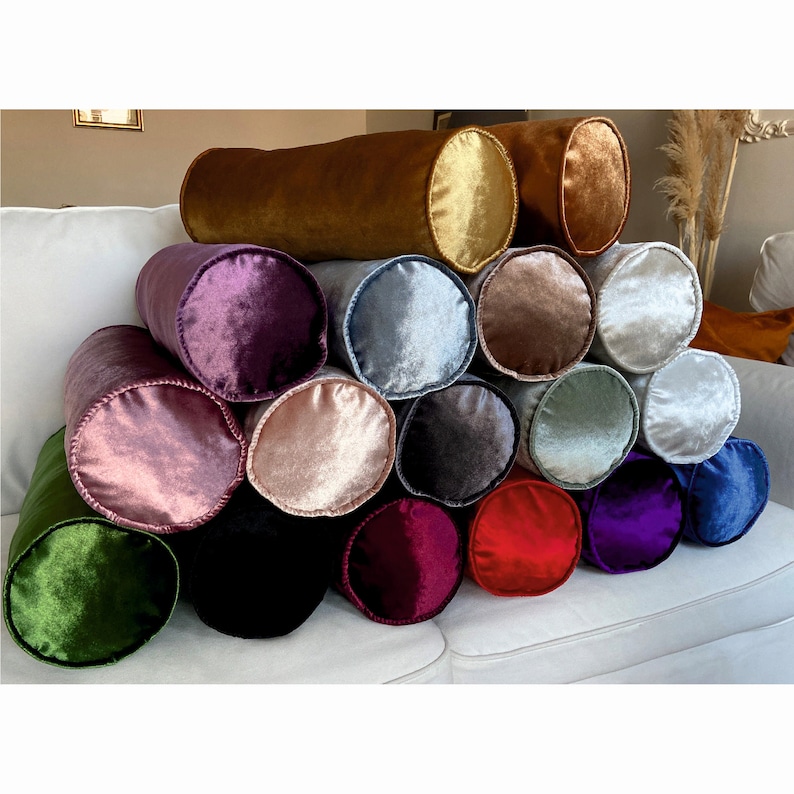 K&ouml;nnte beinhalten: Ein Stapel von 16 zylindrischen Samtkissen in verschiedenen Farben, darunter Gr&uuml;n, Lila, Rosa, Rot und Blau. Die Kissen sind pyramidenf&ouml;rmig auf einem wei&szlig;en Sofa angeordnet.
