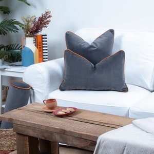 May include: Two gray velvet throw pillows with orange trim on a white couch. The pillows are arranged on a white couch with a wooden coffee table in front of it. The coffee table has a small red dish with cookies on it and a small brown cup and saucer.