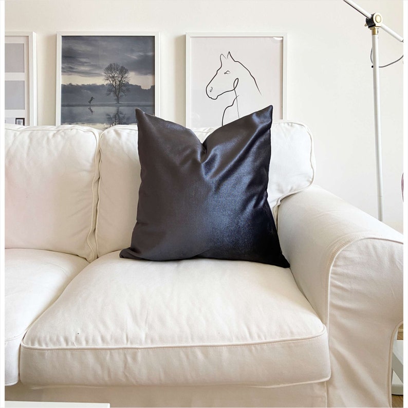 May include: A white couch with a dark gray velvet pillow. The couch is in a living room with white walls and framed artwork.