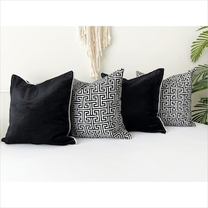 May include: Four decorative throw pillows, two black velvet and two with a white and black geometric pattern. The pillows are arranged on a white couch.