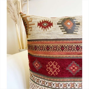 May include: A decorative throw pillow with a geometric pattern in shades of red, orange, brown, and beige. The pillow has a woven texture and a fringe border.