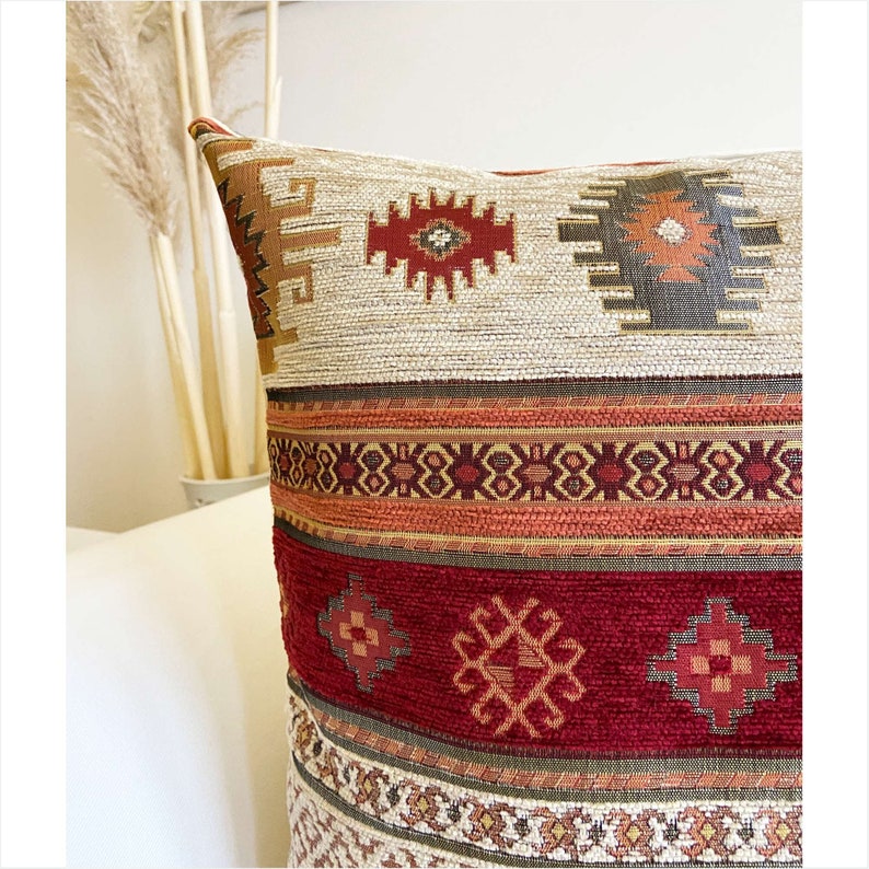 May include: A decorative throw pillow with a geometric pattern in shades of red, orange, brown, and beige. The pillow has a woven texture and a fringe border.