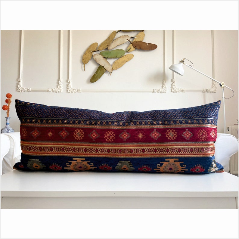 May include: A long, rectangular pillow with a blue, red, and gold geometric pattern. The pillow is made of a soft, textured fabric and has a fringe trim.