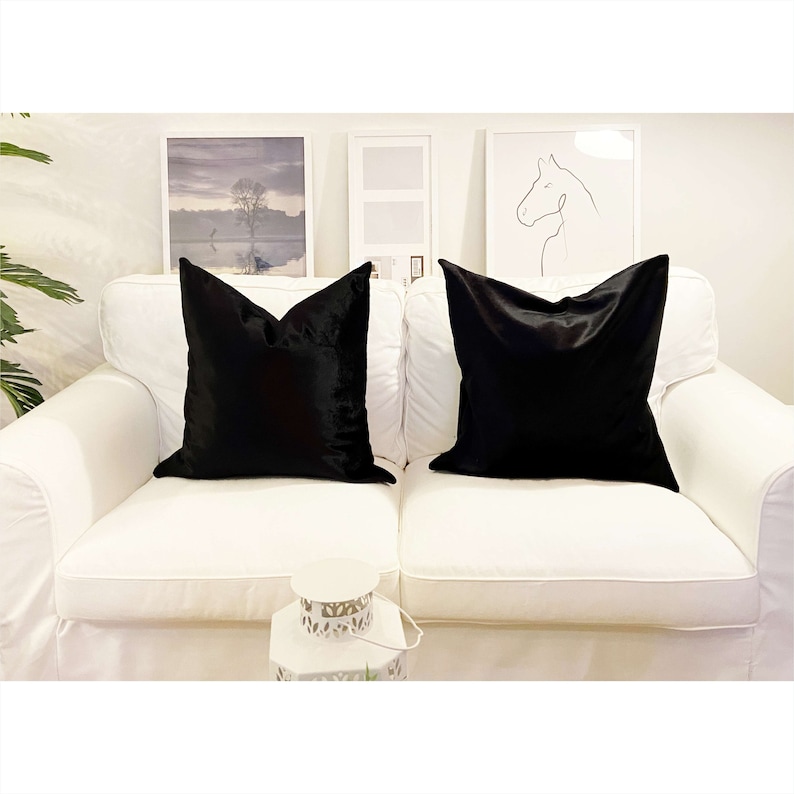May include: Two black velvet throw pillows on a white couch. The pillows are square and have a smooth, soft texture.