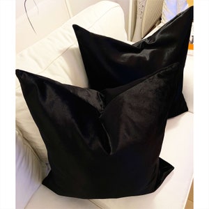 May include: Two black velvet throw pillows on a white couch. The pillows are square and have a smooth, luxurious texture.