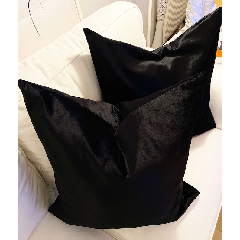 May include: Two black velvet throw pillows on a white couch. The pillows are square and have a smooth, luxurious texture.