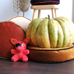 May include: A red, round velvet cushion with a green trim, a tan round velvet cushion with a black trim, and a large green pumpkin on top of a tan round velvet cushion with a black trim. A red plush toy with a black eye patch is in front of the cushions.
