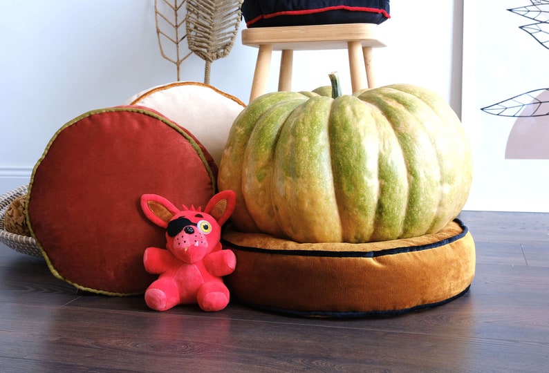 May include: A red, round velvet cushion with a green trim, a tan round velvet cushion with a black trim, and a large green pumpkin on top of a tan round velvet cushion with a black trim. A red plush toy with a black eye patch is in front of the cushions.