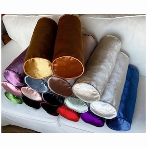 K&ouml;nnte beinhalten: Eine Sammlung von 15 zylindrischen Samt-Kissen in verschiedenen Farben, darunter Gold, Braun, Lila, Blau, Gr&uuml;n und Rosa. Die Kissen sind in einem Halbkreis auf einem wei&szlig;en Sofa angeordnet.